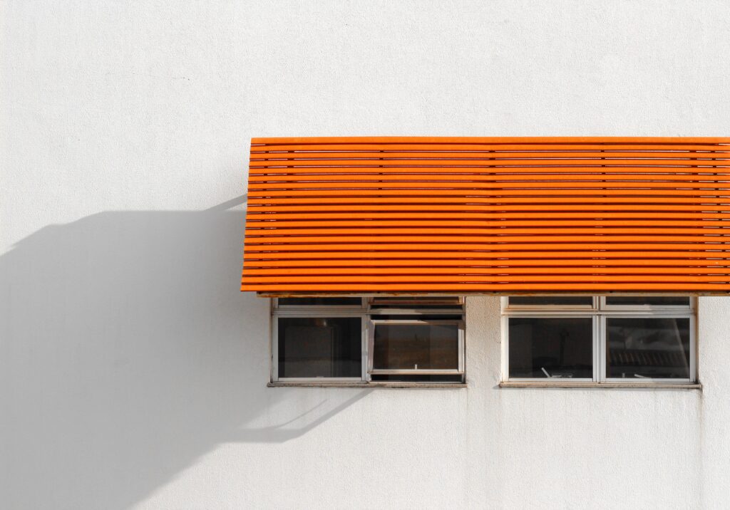 Um toldo laranja moderno projeta sombras em uma fachada de edifício branca com janelas, evidenciando a psicologia do laranja como um ponto focal dinâmico que suaviza a entrada da luz tropical e protege o espaço.