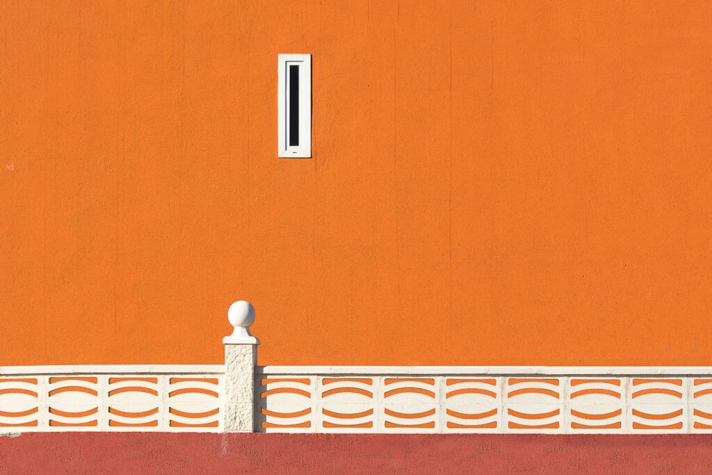 Minimalist orange wall with white patterned railing and a small window in urban setting.