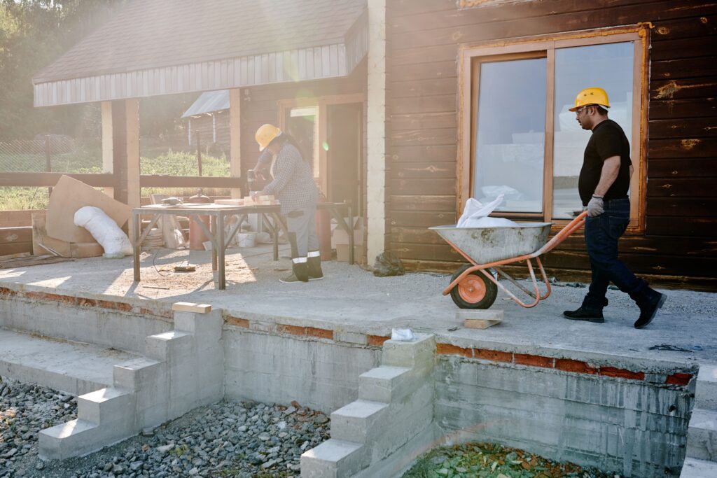 Dois trabalhadores da construção civil realizando reformas na fachada de uma casa, carregando materiais e trabalhando com madeira, ideal para ilustrar uma calculadora de custos de construção.